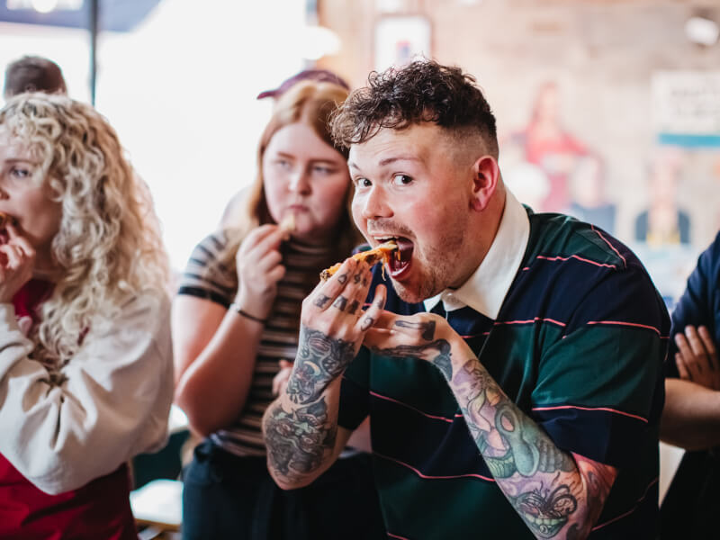 A man is standing in a group of people, about to take a bite of a slice of handmade pizza